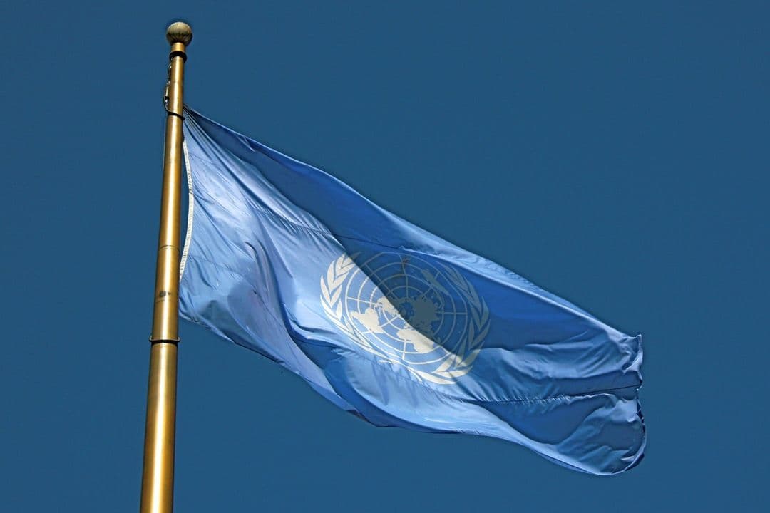 Light blue United Nations flag waving on a gold pole against a deep blue sky