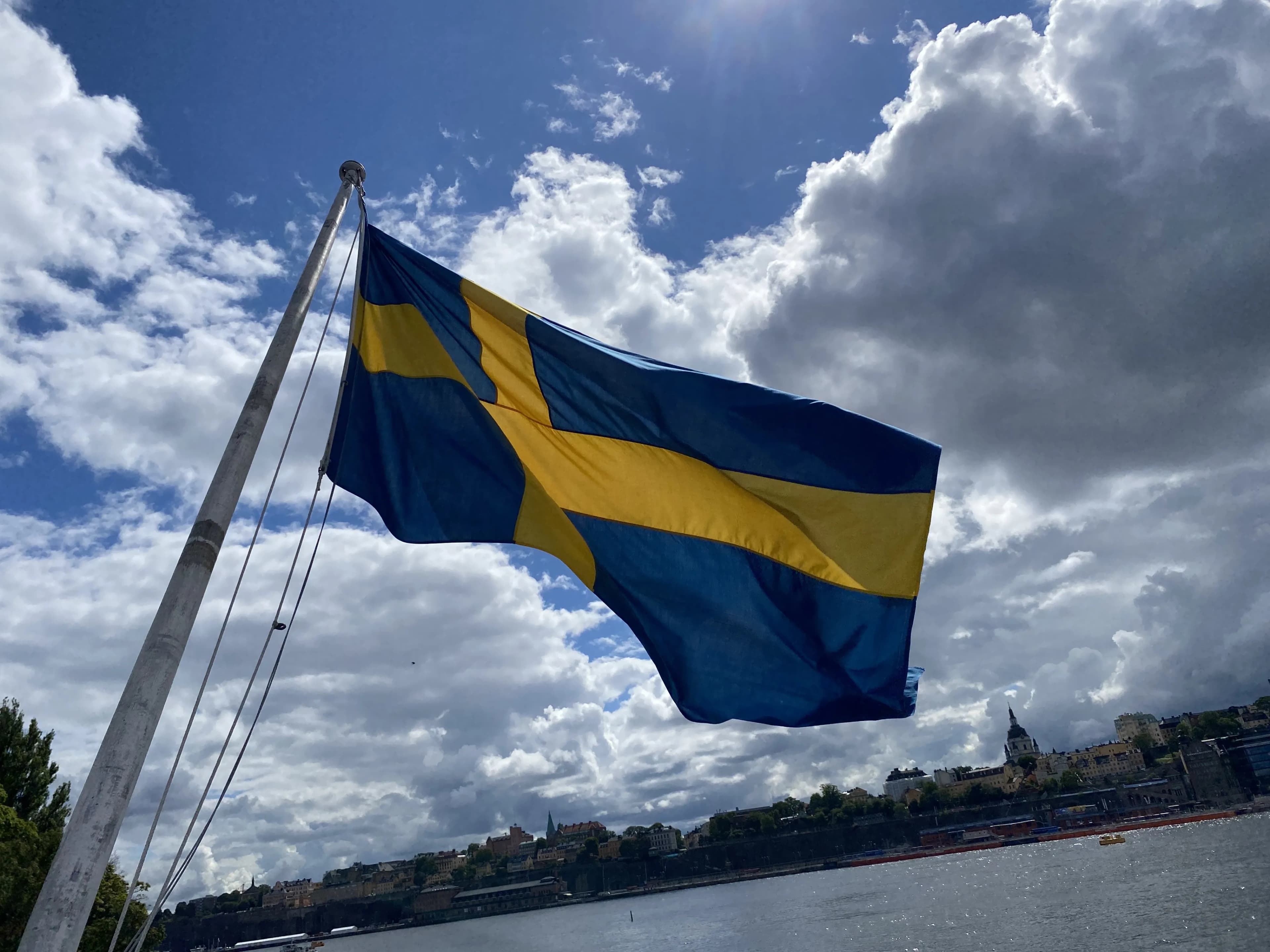 The Swedish flag flying from a mast over Stockholm's water on a bright, cloudy day