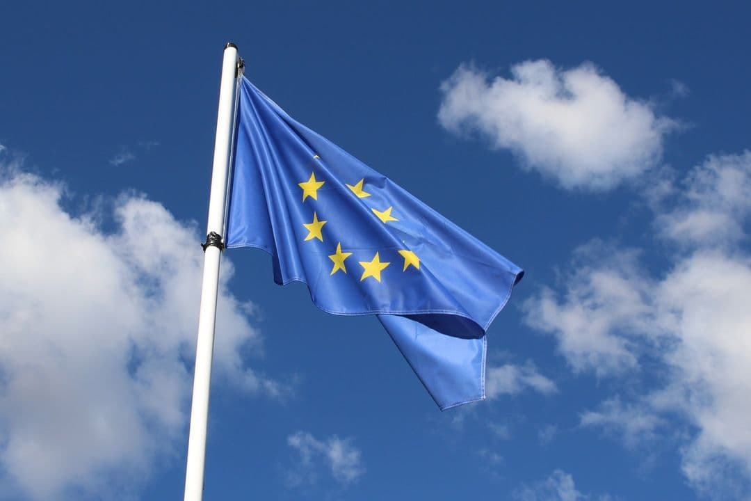 EU flag with circle of gold stars waving on a white pole against blue sky
