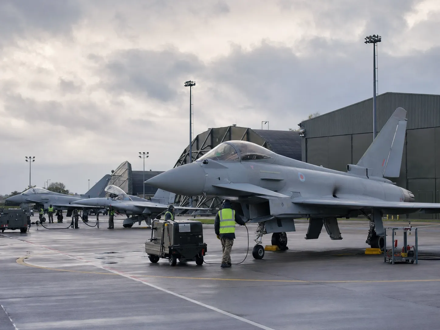 RAF Typhoon fighter jets on a wet flightline as ground crew service them beside a hangar
